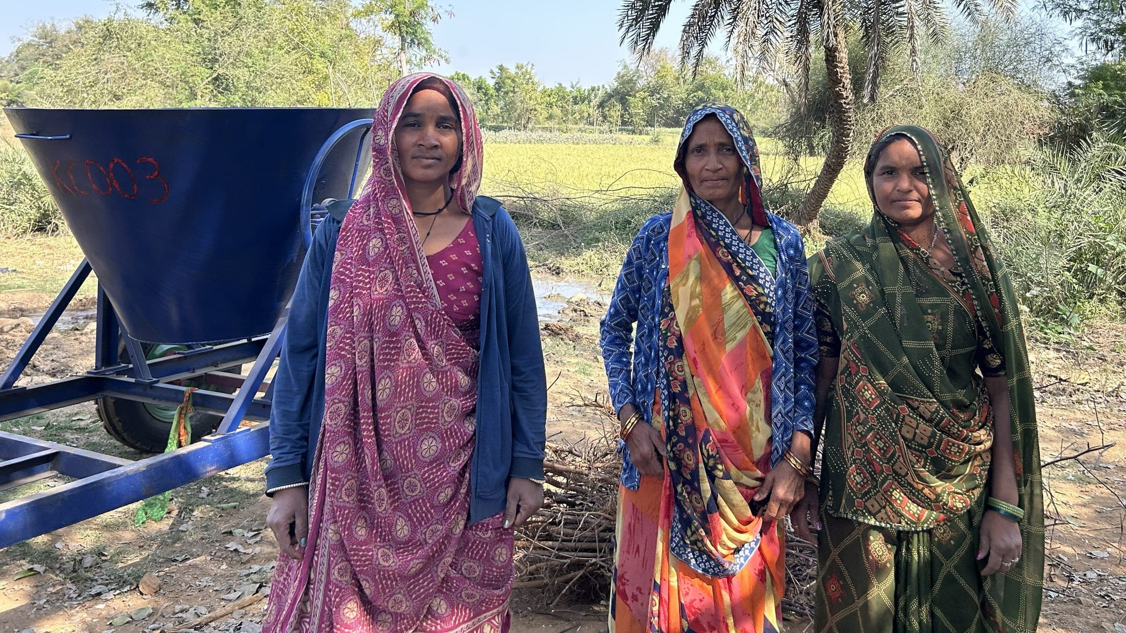 Three women farmers standing beside a biochar kiln at the edge of a field