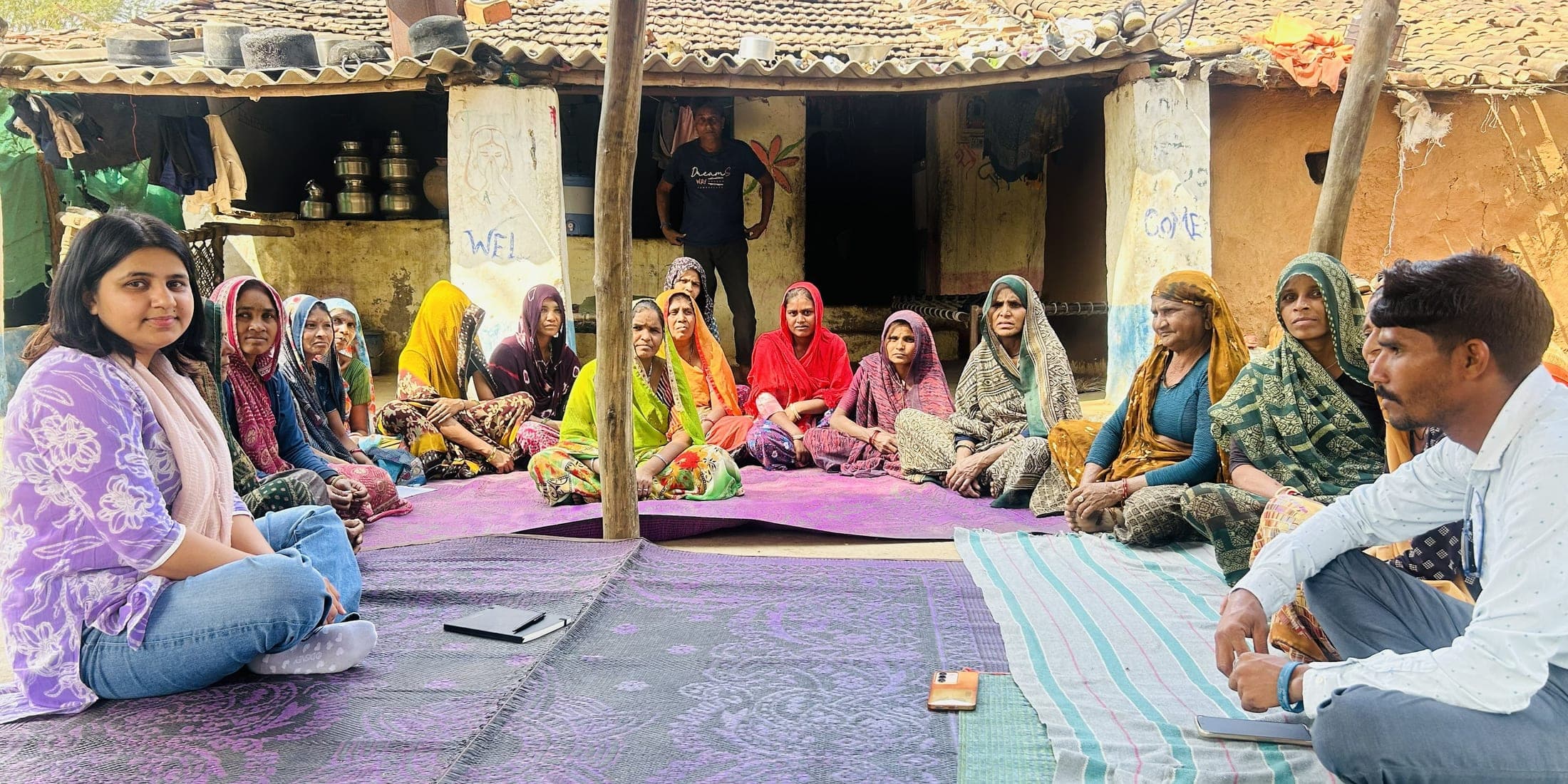 Women farmers sit in a circle on mats in the yard of a village house, with two visitors in the foreground
